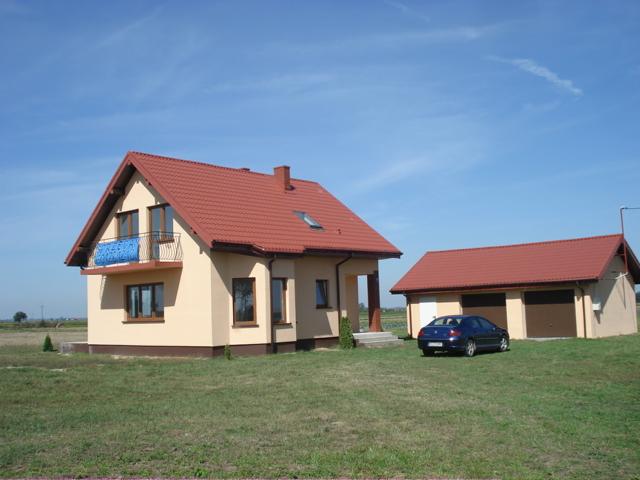 Nowo wybudowany dom jednorodzinny z garażem wolnostojącym, beżowa elewacja, czerwony dach, niebieski samochód na trawniku, błękitne niebo w tle.