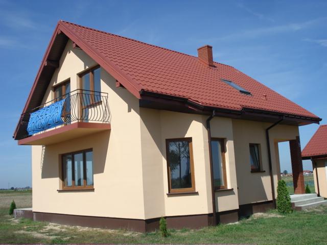 Beżowy dom jednorodzinny z czerwonym dachem i balkonem z niebieską reklamą, widok z zewnątrz w słoneczny dzień.