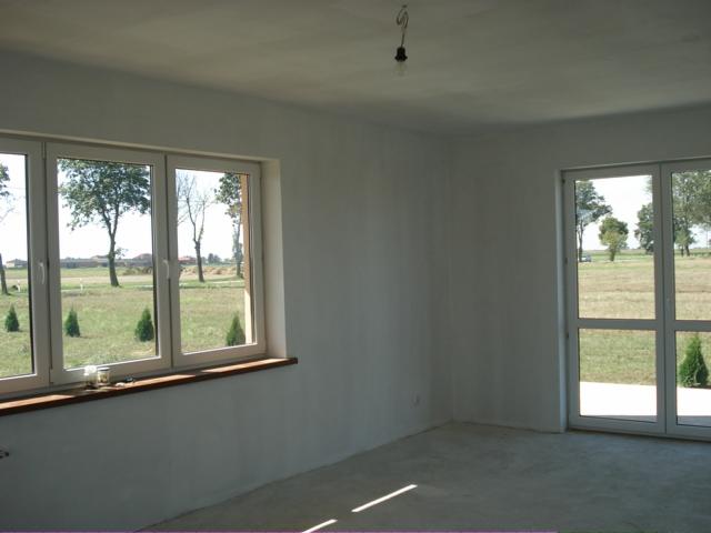 Surowe wnętrze pokoju z białymi ścianami i dużymi oknami z widokiem na zielony krajobraz, widoczny surowy beton na podłodze i wisząca żarówka bez klosza.