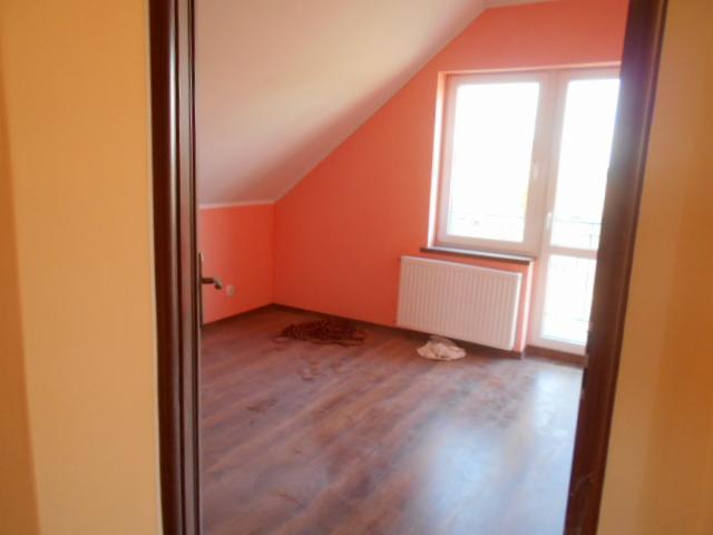 Poddasze z oknem dachowym, pomalowane na brzoskwiniowo, z drewnianą podłogą, widziane z perspektywy drzwi, z widocznym grzejnikiem pod oknem i zwiniętym kablem na podłodze.