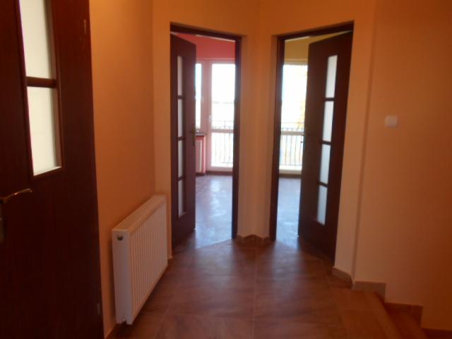 Korytarz w odcieniach beżu z brązowymi drzwiami z przeszkleniami, widoczne dwa otwarte wejścia do pokojów z balkonami, na ścianie biały grzejnik.