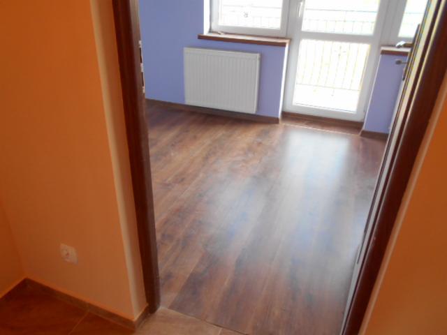 Widok z perspektywy korytarza na pokój z panelami podłogowymi w odcieniach brązu, białym grzejnikiem pod oknem i fioletowymi ścianami.