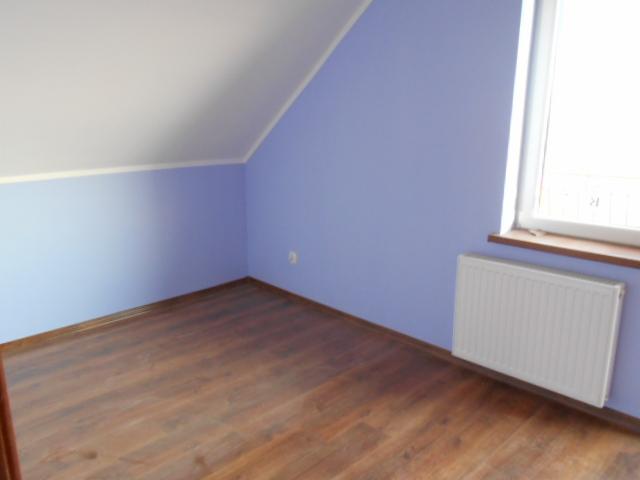 Pusty pokój na poddaszu z panelami podłogowymi w odcieniach brązu, błękitnymi ścianami, białym sufitem ze skosem i białym grzejnikiem pod oknem.