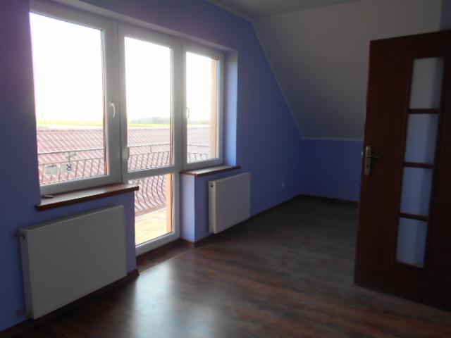 Pusty pokój na poddaszu z oknem balkonowym, grzejnikami i drewnianą podłogą w odcieniach brązu, ściany pomalowane na fioletowo, widok na balkon i okolicę.