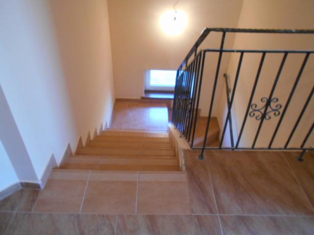 Klatka schodowa wykończona brązowymi płytkami, z czarną, metalową balustradą z ozdobnym motywem, widziana z góry w kierunku okna na półpiętrze.