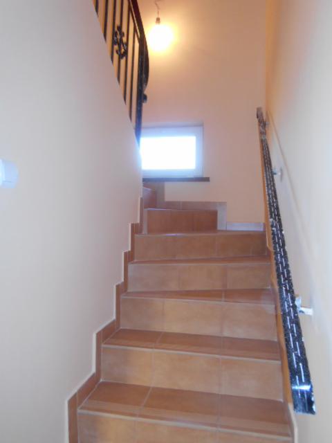 Schody wewnętrzne wyłożone brązowymi płytkami ceramicznymi, z kutą, czarną balustradą i widocznym oknem na półpiętrze, oświetlone górną lampą.