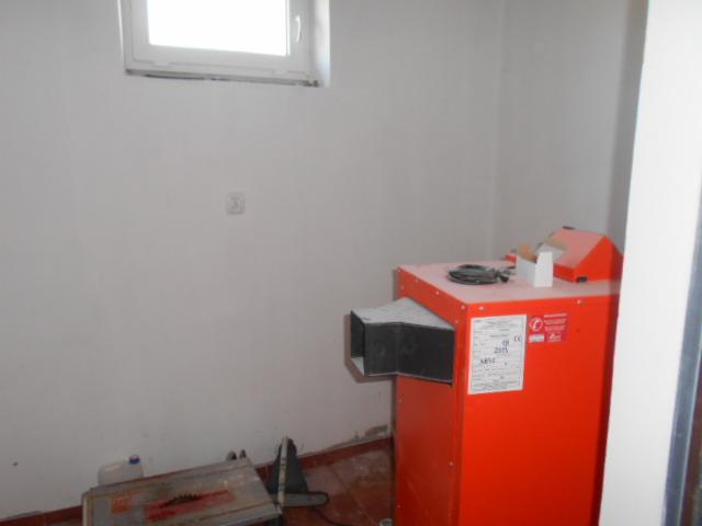 Pomarańczowy piec centralnego ogrzewania w rogu pomieszczenia, widoczna piła tarczowa i okno w tle.