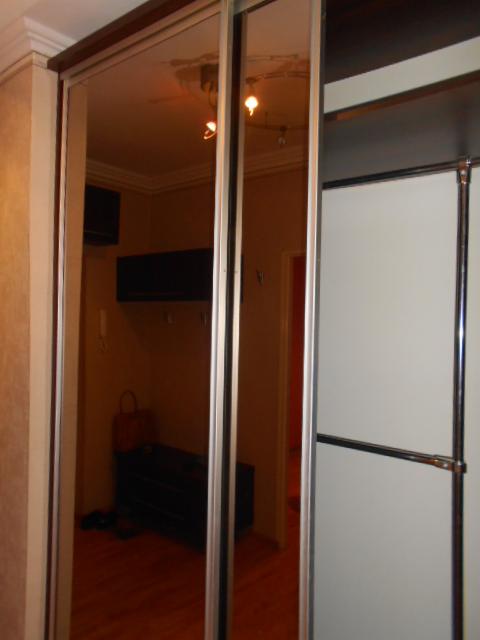 Wnętrze pokoju odbite w lustrzanych drzwiach szafy przesuwnej z aluminiowymi ramami, widoczny fragment białej szafy po prawej stronie.