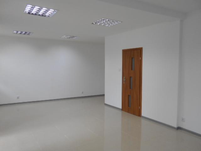 Puste, nowo wyremontowane pomieszczenie biurowe z drewnianymi drzwiami i błyszczącą podłogą z płytek, oświetlone przez rastrowe lampy sufitowe.