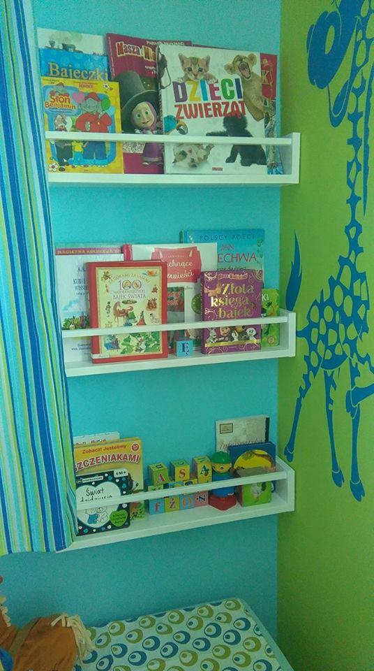 Trzy białe półki z książkami i zabawkami dla dzieci na turkusowej ścianie, obok zielonej ściany z niebieską żyrafą. Na półkach widoczne książki, m.in. 'Bajeczki', 'Dzieci Zwierząt', '100 Baiek...