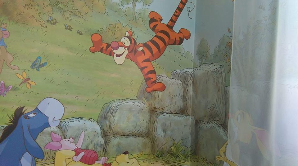 Malowidło ścienne w pokoju dziecięcym przedstawiające postacie z bajki Kubuś Puchatek: Tygrysek skaczący po kamieniach, Kłapouchy i Prosiaczek leżący obok siebie, w tle łąka z motylami.