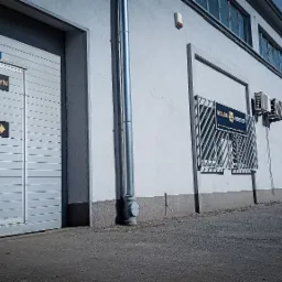 Zewnętrzny widok magazynu z szarymi drzwiami rolowanymi, oznaczonymi tabliczkami 'Magazyn' i 'Biuro', oraz tablicą reklamową 'Yellow Monster' na ścianie budynku.