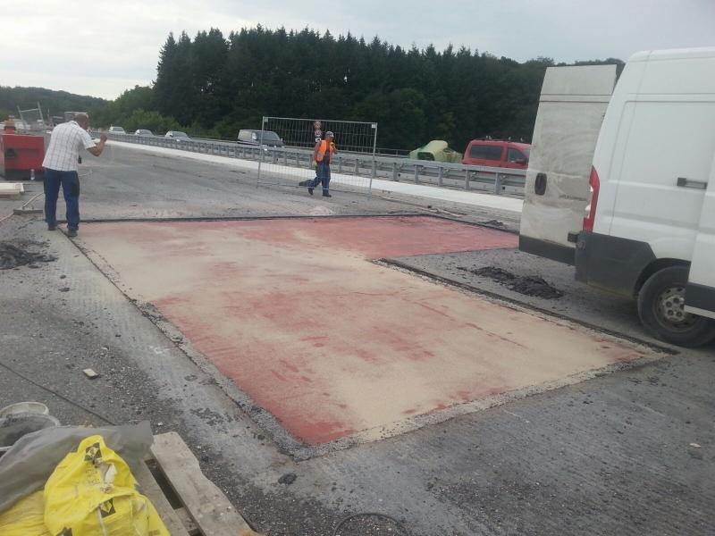 Przygotowanie podłoża na moście, widoczne dwie sekcje z czerwoną warstwą gruntującą, pracownik robiący zdjęcie i drugi w kamizelce odblaskowej przechodzący obok, biały samochód dostawczy w tle.