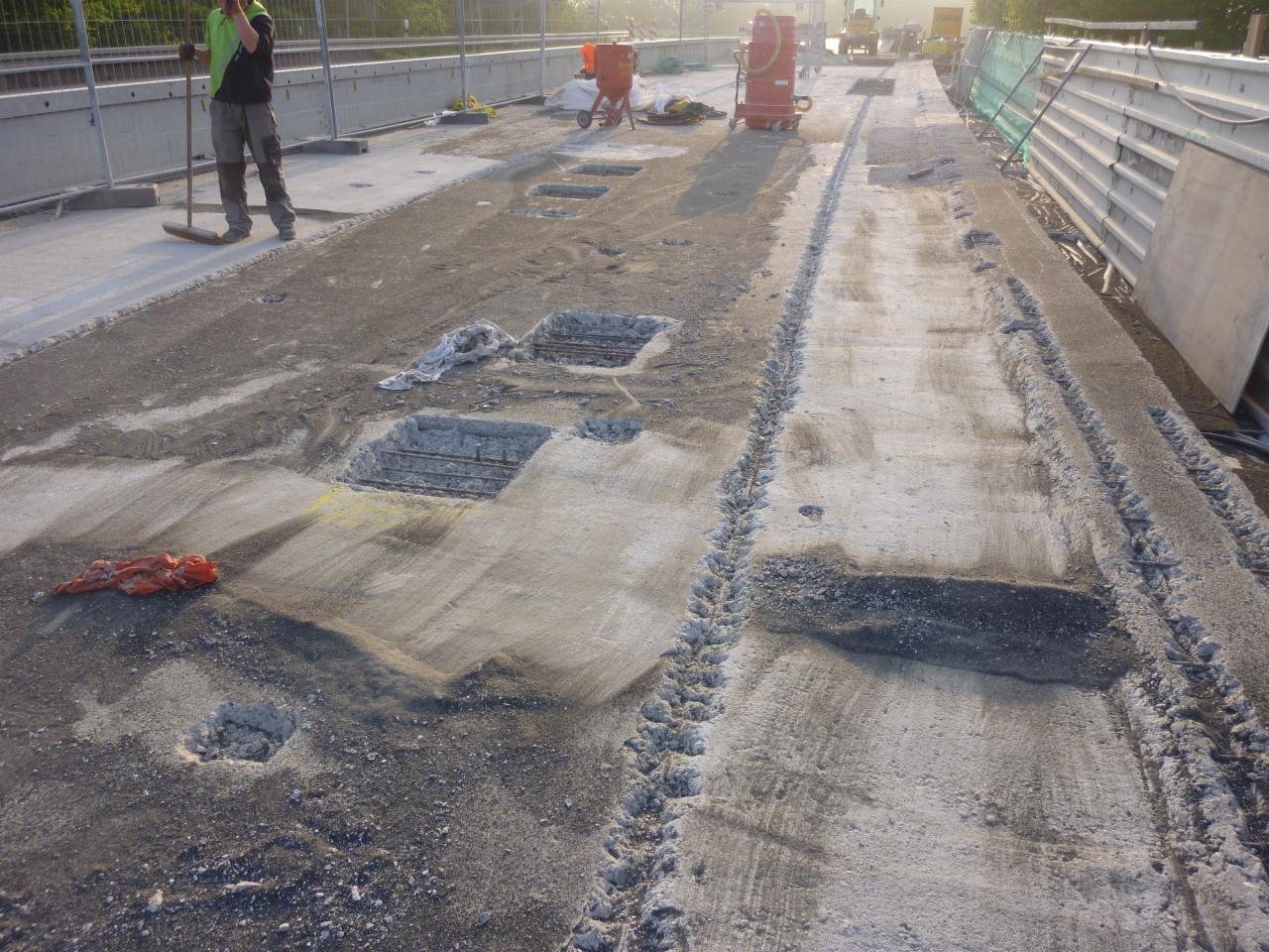 Przygotowanie podłoża pod posadzkę jastrychową: widoczne wycięte fragmenty betonu z odsłoniętym zbrojeniem, ślady opon i pracownik z miotłą w tle.