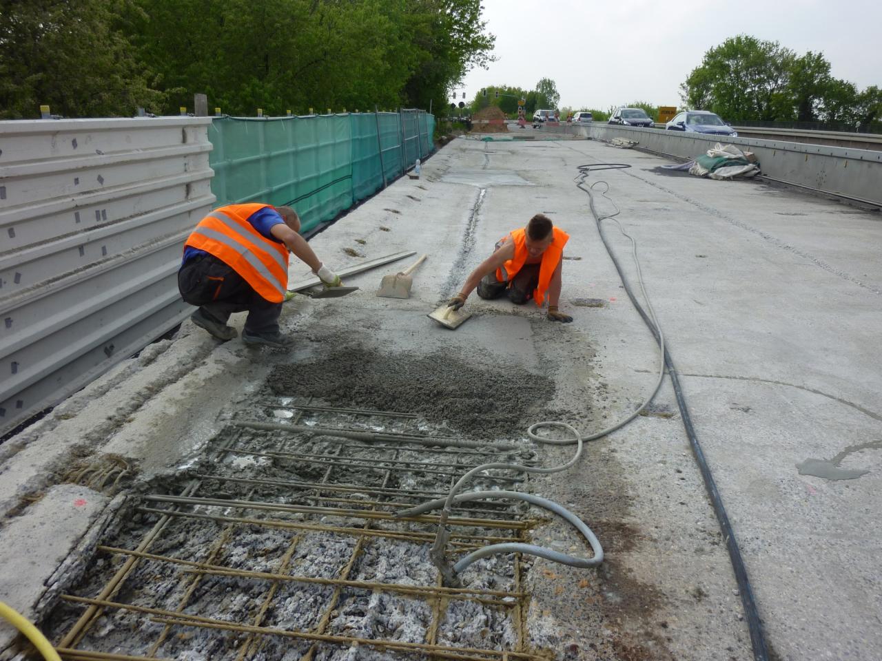 Dwóch pracowników w pomarańczowych kamizelkach wyrównuje świeży beton na powierzchni zbrojonej siatką stalową, prawdopodobnie podczas naprawy mostu lub wiaduktu.