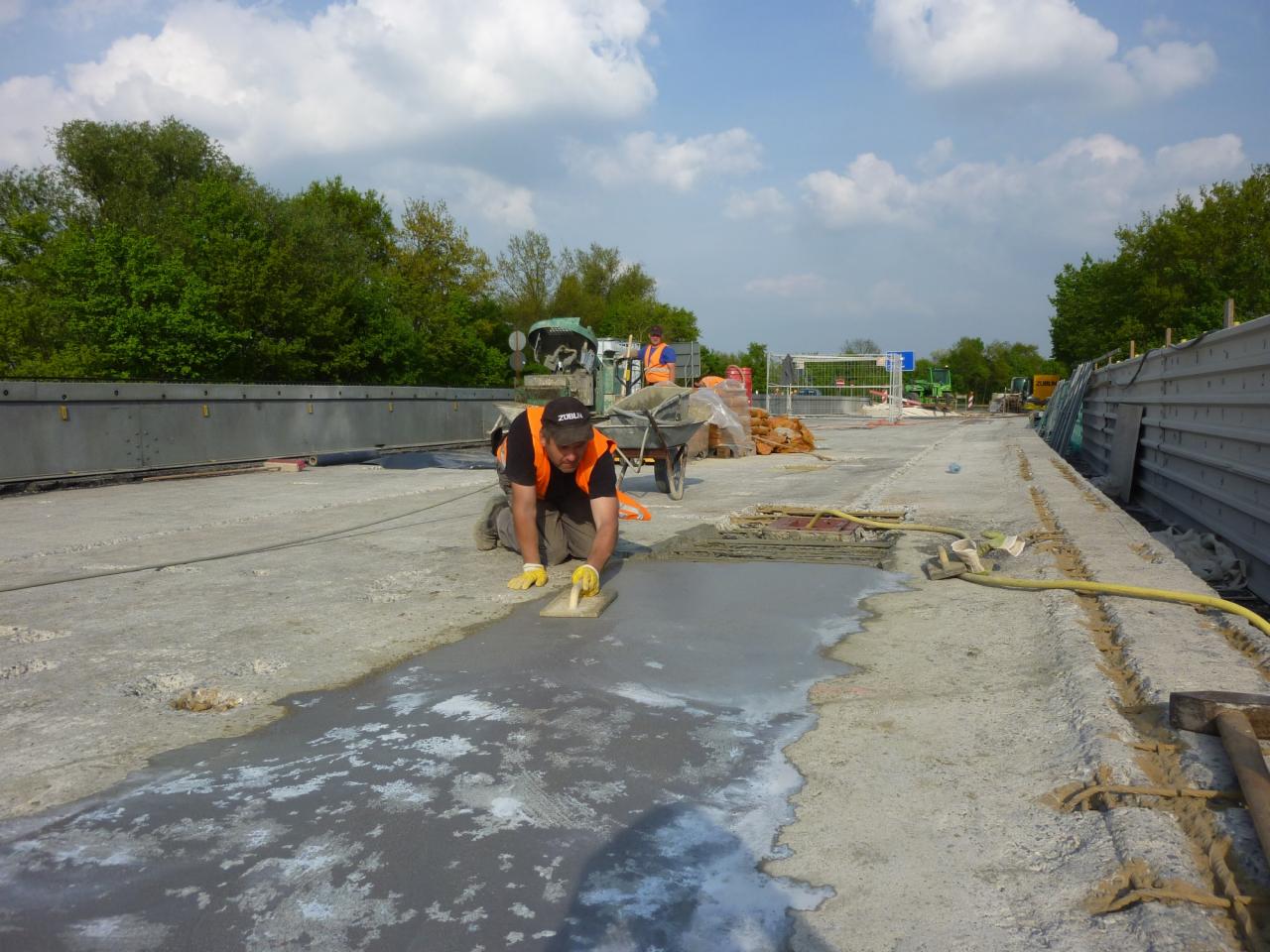 Pracownik w pomarańczowej kamizelce wyrównuje świeżo wylaną betonową posadzkę na wiadukcie przy użyciu packi, w tle widoczne maszyny budowlane i drzewa.