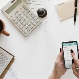 Widok z góry na białe biurko z kalkulatorem, kawą, gazetą biznesową, telefonem wyświetlającym aplikację 'Faktury' obsługiwaną przez ręce z ciemnoczerwonym manicure i dokumentami, sugerujący pracę...