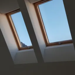 Dwa okna dachowe z drewnianą ramą, widok na błękitne niebo, ściany pomalowane na biało, delikatne cieniowanie światła.