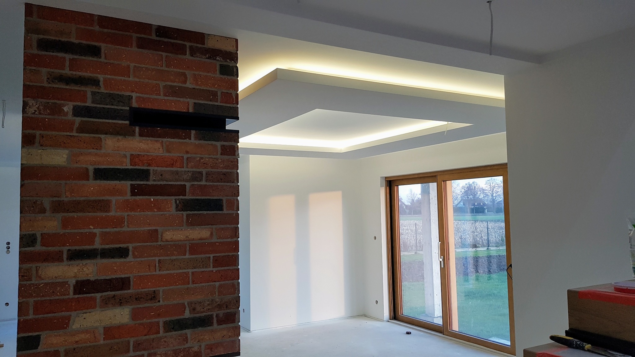 Nowoczesne wnętrze z ceglaną ścianą i podwieszanym sufitem z oświetleniem LED, widok na ogród przez przesuwne okno.