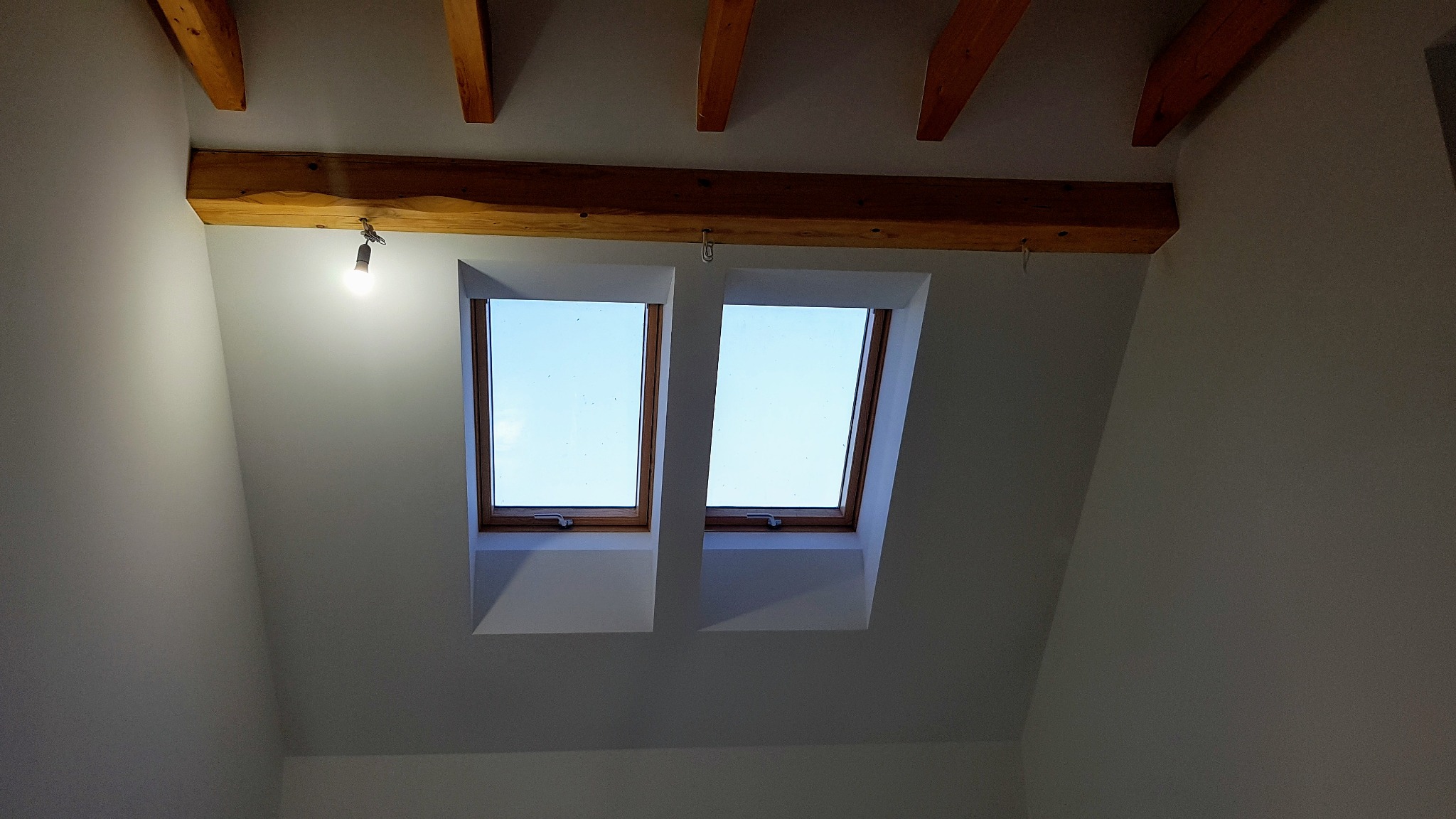 Świeżo pomalowane poddasze ze skośnymi ścianami, drewnianymi belkami stropowymi i dwoma oknami dachowymi, oświetlone wiszącą żarówką.
