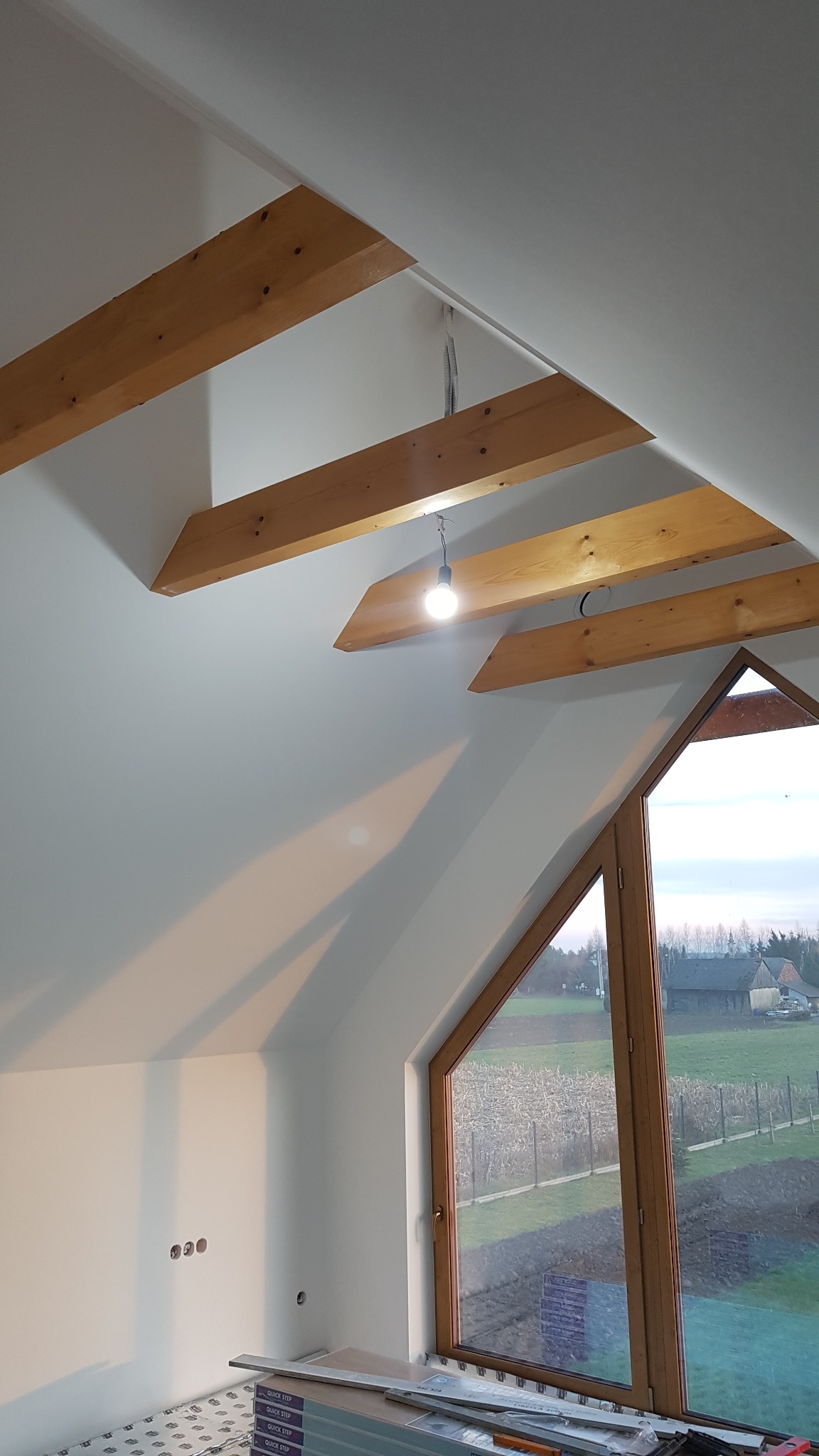 Poddasze po remoncie z drewnianymi belkami stropowymi, świeżo pomalowanymi ścianami i oknem z widokiem na zielony krajobraz. Na podłodze widoczne panele podłogowe przed montażem.