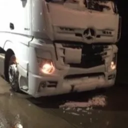 Biała ciężarówka Mercedes-Benz Actros z przodu, pokryta śniegiem, stojąca na mokrej nawierzchni w nocy, z włączonymi światłami.