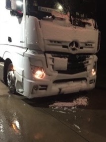 Biała ciężarówka Mercedes-Benz Actros z przodu, pokryta śniegiem, stojąca na mokrej nawierzchni w nocy, z włączonymi światłami.