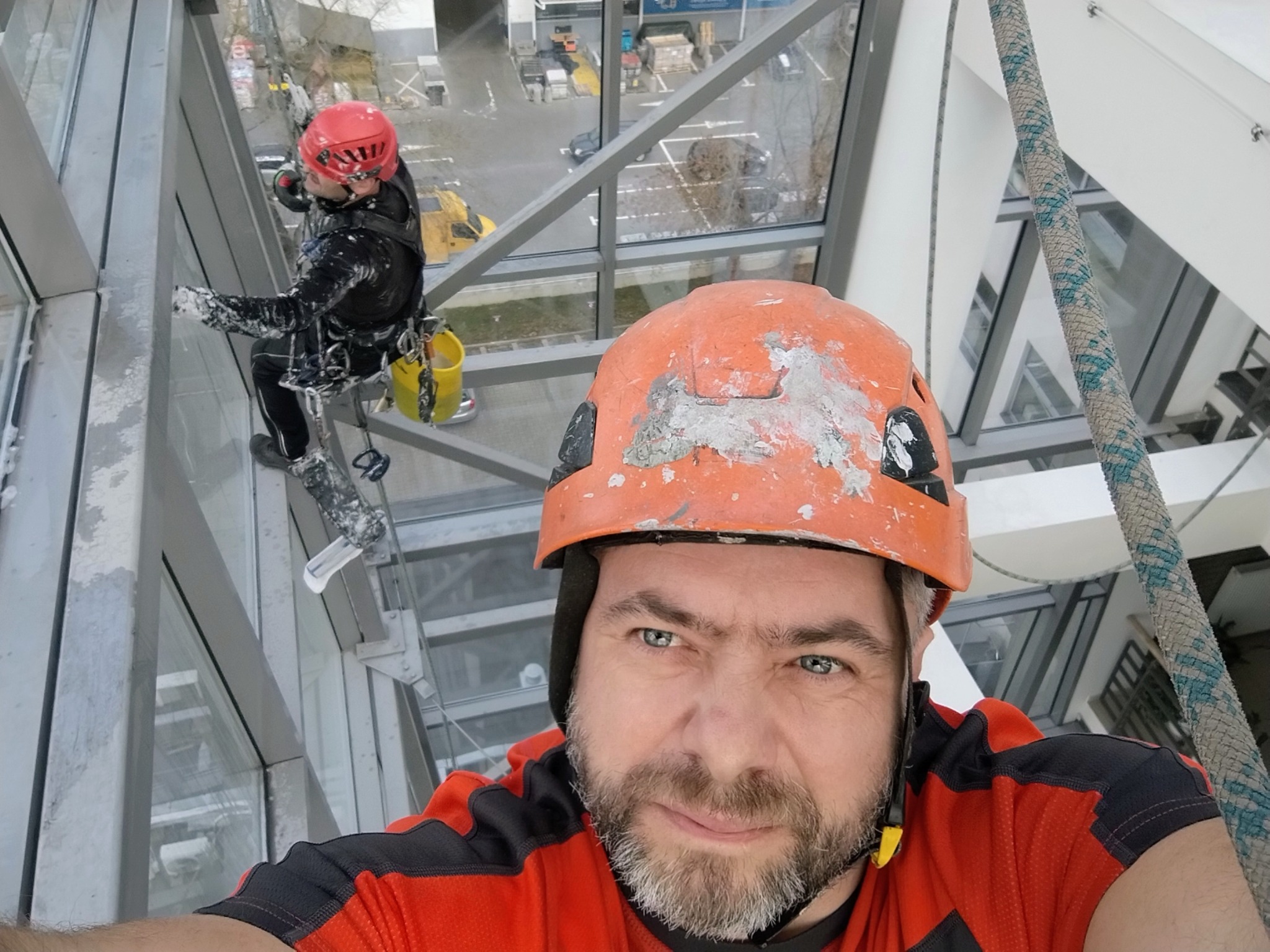 Pracownik w kasku, na linach, myje okna wieżowca. Widok z dołu, drugi pracownik w tle. Praca na wysokościach z użyciem technik alpinistycznych.