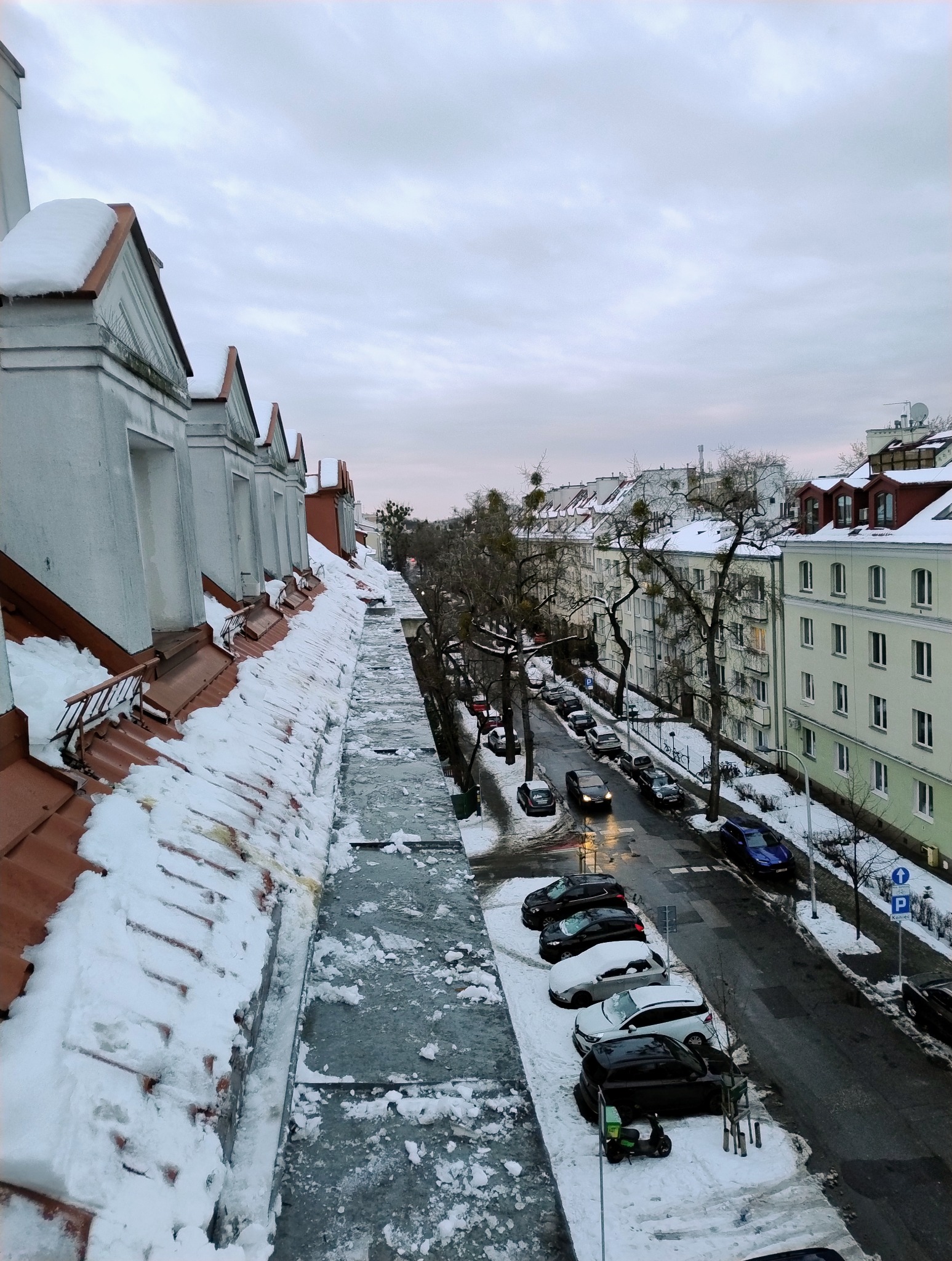 Śnieg na dachu budynku z widokiem na ulicę i zaparkowane samochody w zimowy dzień. Widok z góry, pochmurne niebo.