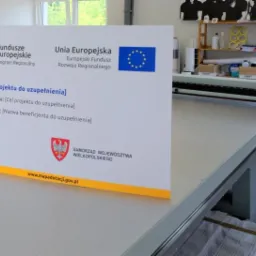 Tablica informacyjna projektu współfinansowanego z funduszy europejskich, umieszczona obok plotera tnącego w drukarni, z widocznym logo Samorządu Województwa Wielkopolskiego.