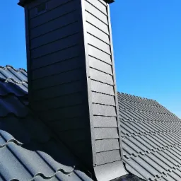 Nowoczesny, grafitowy komin wentylacyjny z blachy na dachu pokrytym grafitową dachówką ceramiczną, widoczne mocowania i obróbki blacharskie, na tle błękitnego nieba.