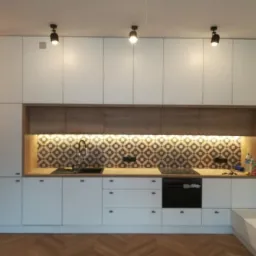 Nowoczesna kuchnia z białymi szafkami i drewnianymi akcentami, oświetlona punktowymi lampami sufitowymi, z dekoracyjną mozaiką nad blatem i parkietem w jodełkę na podłodze.