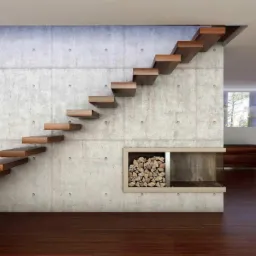 Nowoczesne, wiszące stopnie schodów z ciemnego drewna na tle betonowej ściany, obok wnęka z drewnem kominkowym, widoczny fragment salonu z dużym oknem i logo firmy w prawym dolnym rogu.