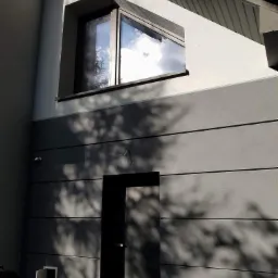 Szara elewacja domu z poziomymi panelami, widoczne okno z odbiciem nieba i ciemne drzwi wejściowe, całość zacieniona przez drzewo.