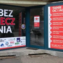 Wejście do biura ubezpieczeń w Gorzowie Wielkopolskim z widocznymi naklejkami informacyjnymi na szybach oraz logo różnych towarzystw ubezpieczeniowych.