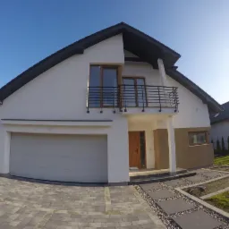 Nowoczesny dom jednorodzinny z białym tynkiem, ciemnym dachem, balkonem z czarną balustradą i szarą bramą garażową, widziany z poziomu kostki brukowej.