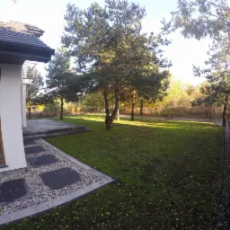 Nowoczesny dom jednorodzinny z zadbanym trawnikiem, ścieżką z kamiennych płyt i żywopłotem z tui wzdłuż ogrodzenia.