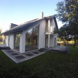 Nowoczesny dom jednorodzinny z dużymi oknami i balkonem, otoczony zielonym trawnikiem i drzewami, z wykończonym tarasem i ścieżką z kamiennych płyt.