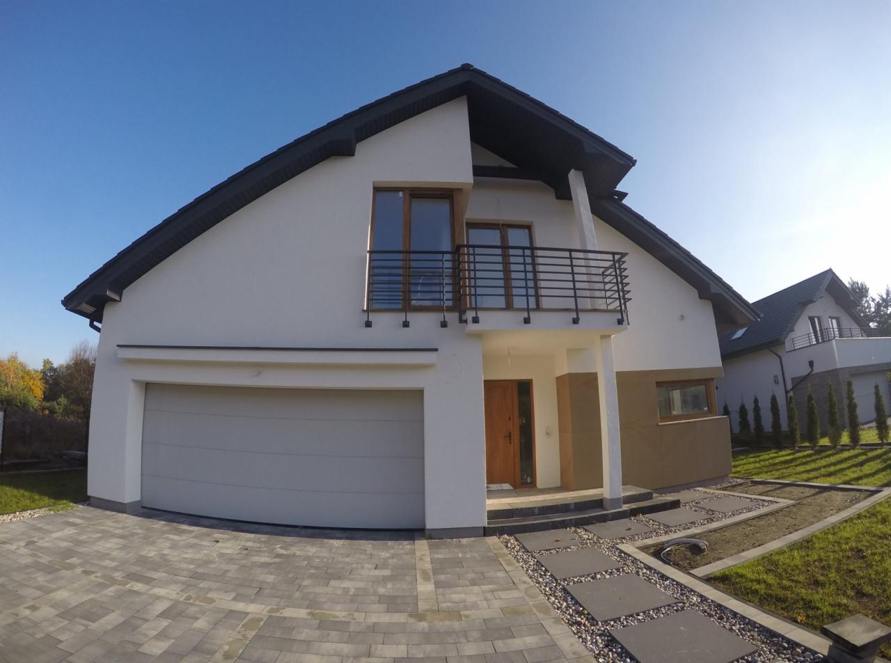 Nowoczesny dom jednorodzinny z białym tynkiem, ciemnym dachem, balkonem z czarną balustradą i szarą bramą garażową, widziany z poziomu kostki brukowej.