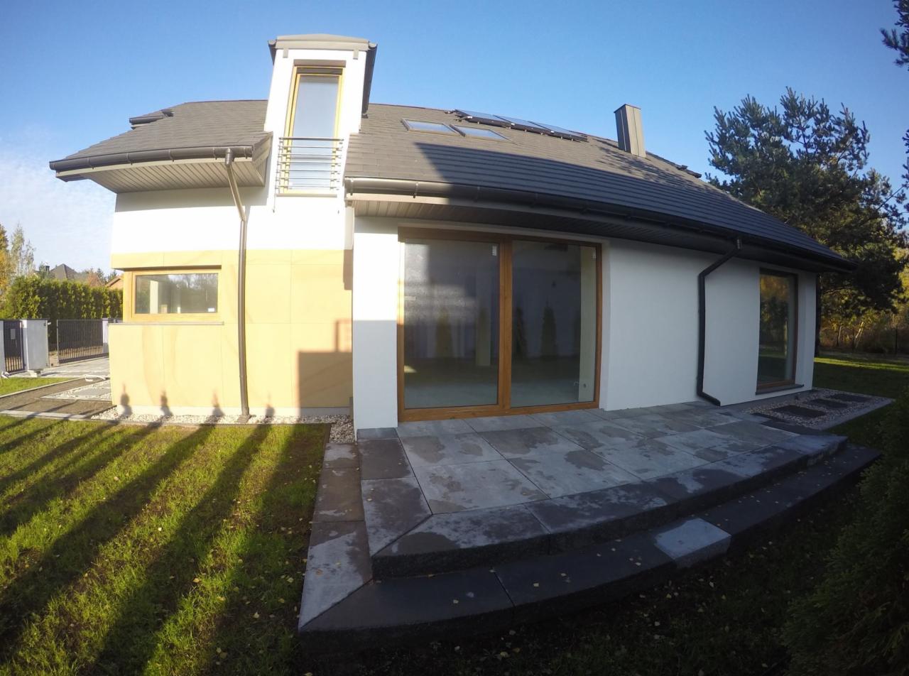 Nowoczesny dom jednorodzinny z elementami wykończenia elewacji w odcieniach beżu i szarości, widoczny taras z kamiennych płyt oraz okna z drewnianą stolarką.