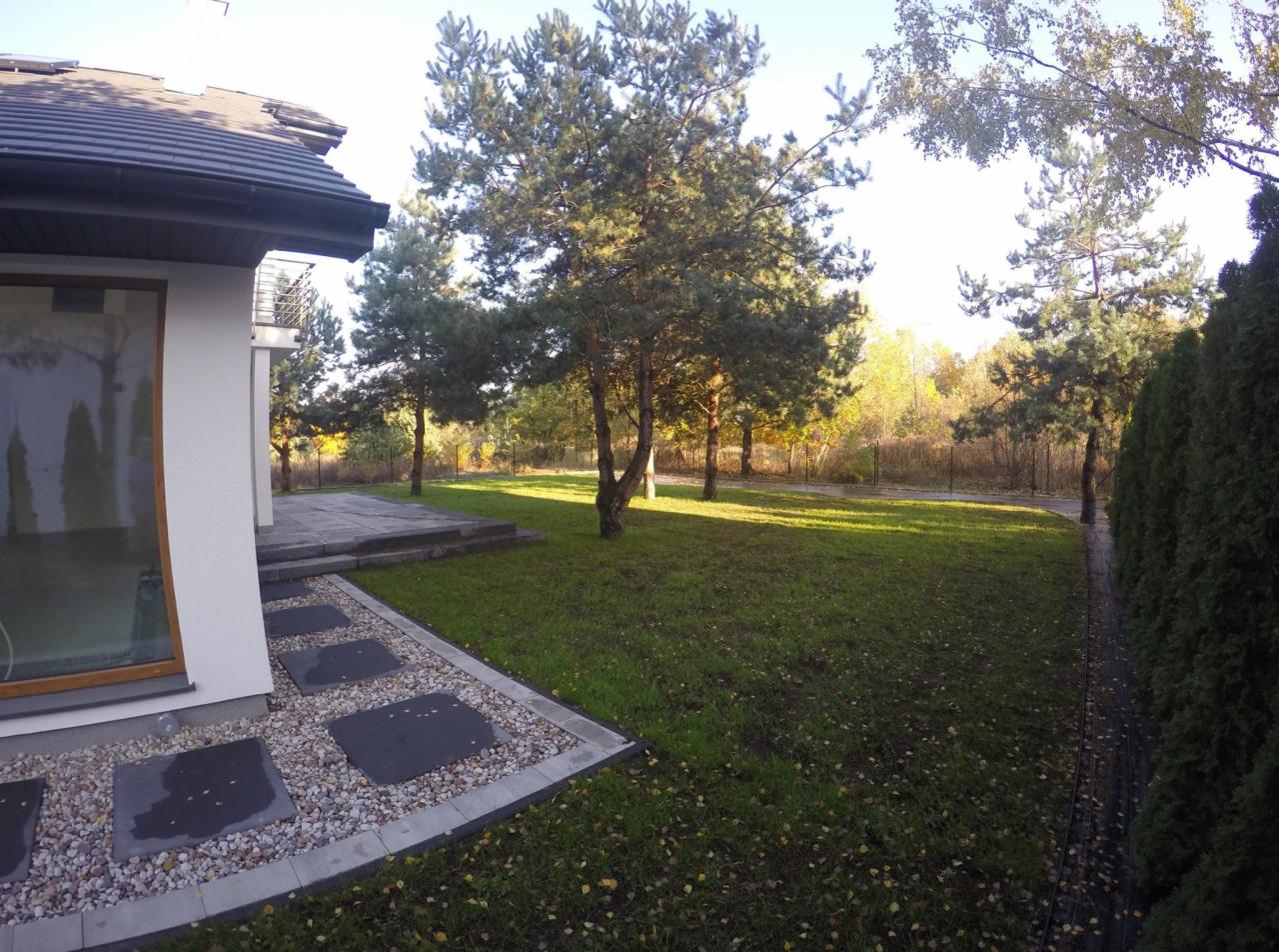 Nowoczesny dom jednorodzinny z zadbanym trawnikiem, ścieżką z kamiennych płyt i żywopłotem z tui wzdłuż ogrodzenia.