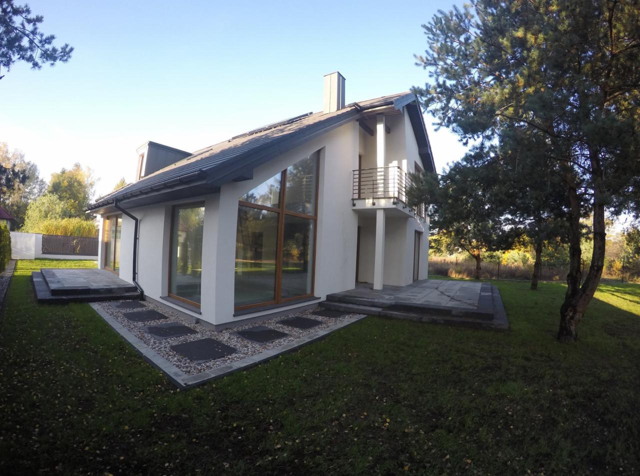 Nowoczesny dom jednorodzinny z dużymi oknami i balkonem, otoczony zielonym trawnikiem i drzewami, z wykończonym tarasem i ścieżką z kamiennych płyt.