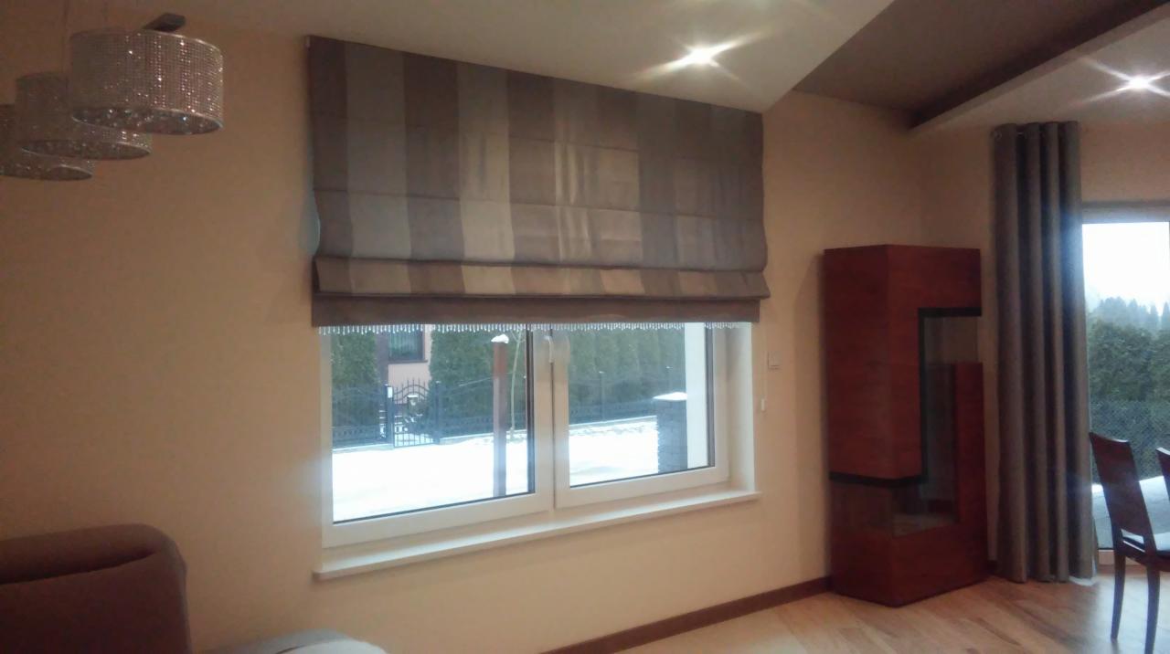 Zasłona rzymska w odcieniach szarości, z widocznymi poziomymi pasami, zamontowana w oknie z widokiem na zimowy krajobraz.