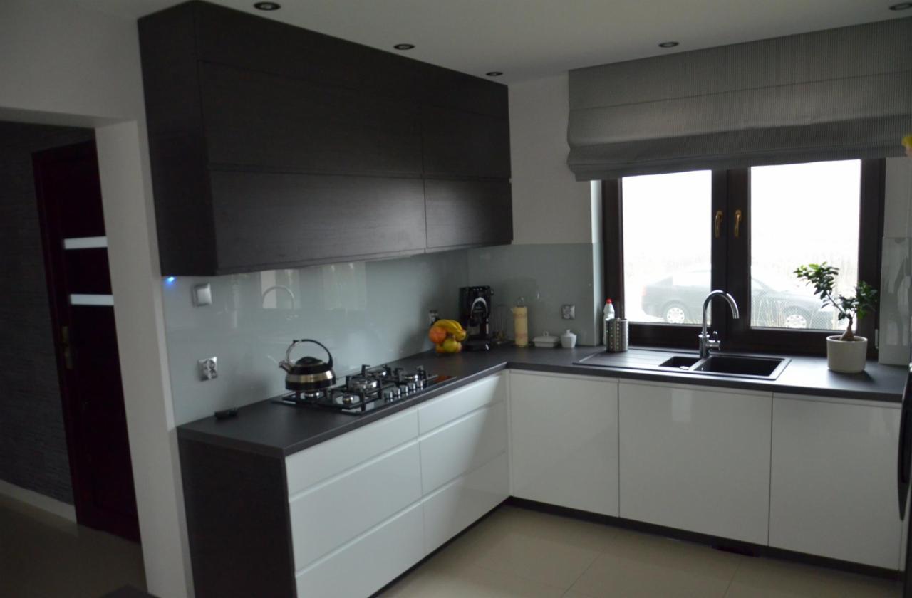 Nowoczesna kuchnia z białymi szafkami i ciemnym blatem, okno z szarą roletą rzymską.