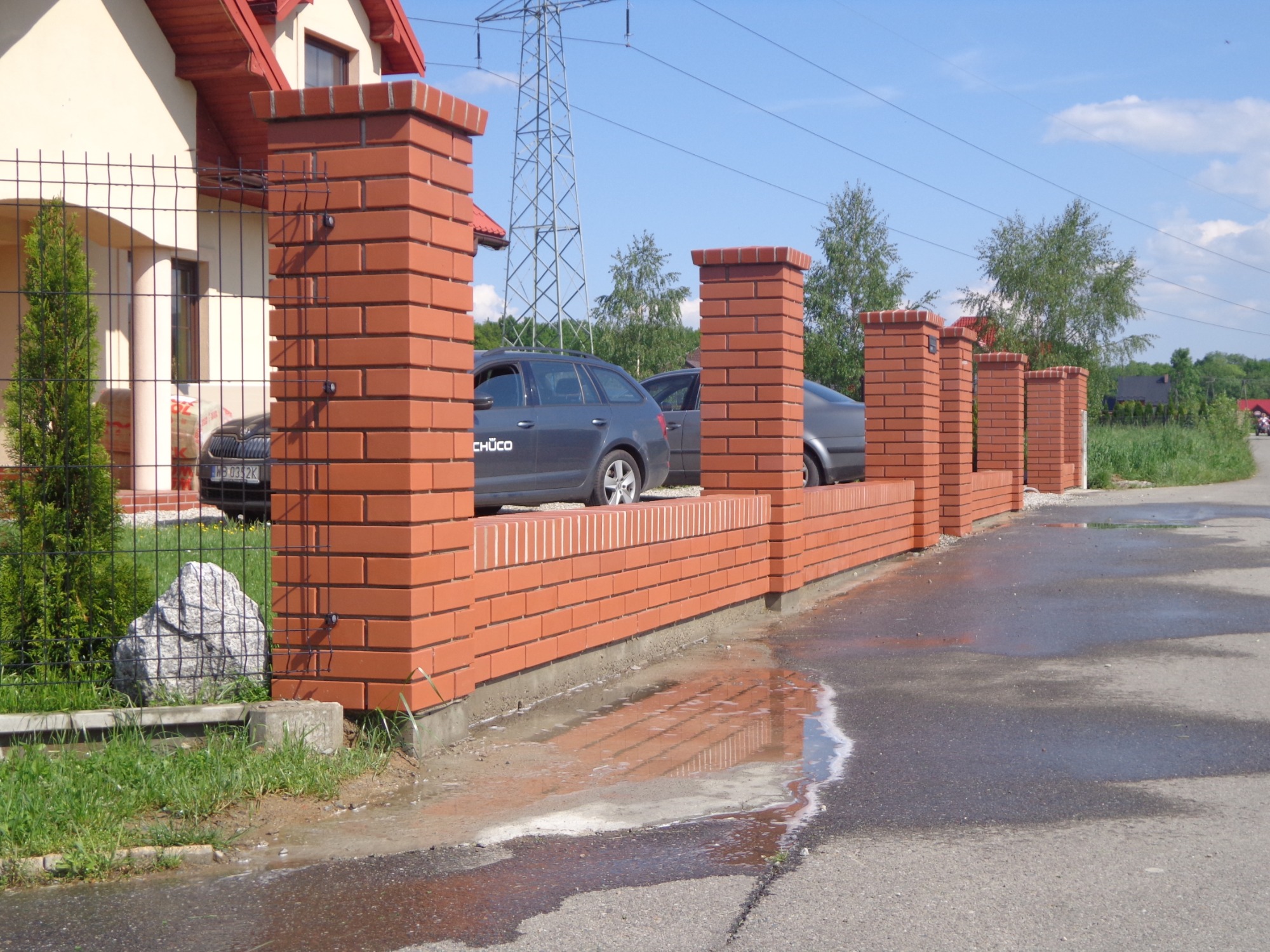 Nowoczesne ogrodzenie z czerwonej cegły klinkierowej z metalową siatką, widok z zewnątrz na ulicę z kałużą po deszczu, w tle zaparkowane samochody i słup wysokiego napięcia.