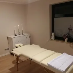 Beżowy gabinet masażu z białym łóżkiem do masażu, ręcznikiem, komodą z zapalonymi świecami i kwiatami w doniczce na parapecie okna.