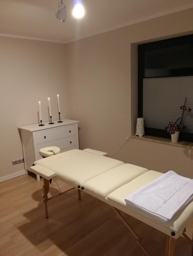 Beżowy gabinet masażu z białym łóżkiem do masażu, ręcznikiem, komodą z zapalonymi świecami i kwiatami w doniczce na parapecie okna.