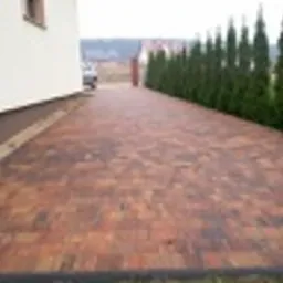 Ułożona kostka brukowa w odcieniach brązu i czerwieni, tworząca wzór na podjeździe przed domem. W tle rząd wysokich, smukłych drzew iglastych i częściowo widoczny budynek z białymi ścianami.