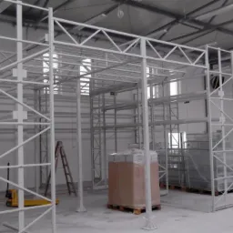 Montaż białych, metalowych regałów magazynowych z widocznymi elementami konstrukcyjnymi, drabina i paleta z towarem w tle, hala magazynowa w trakcie wyposażania.