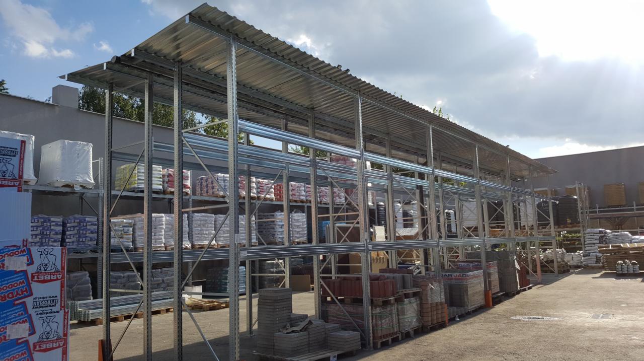 Metalowa konstrukcja regałowa zadaszona blachą falistą, wypełniona materiałami budowlanymi, takimi jak kostka brukowa, worki z zaprawą i styropian, składowana na zewnątrz.